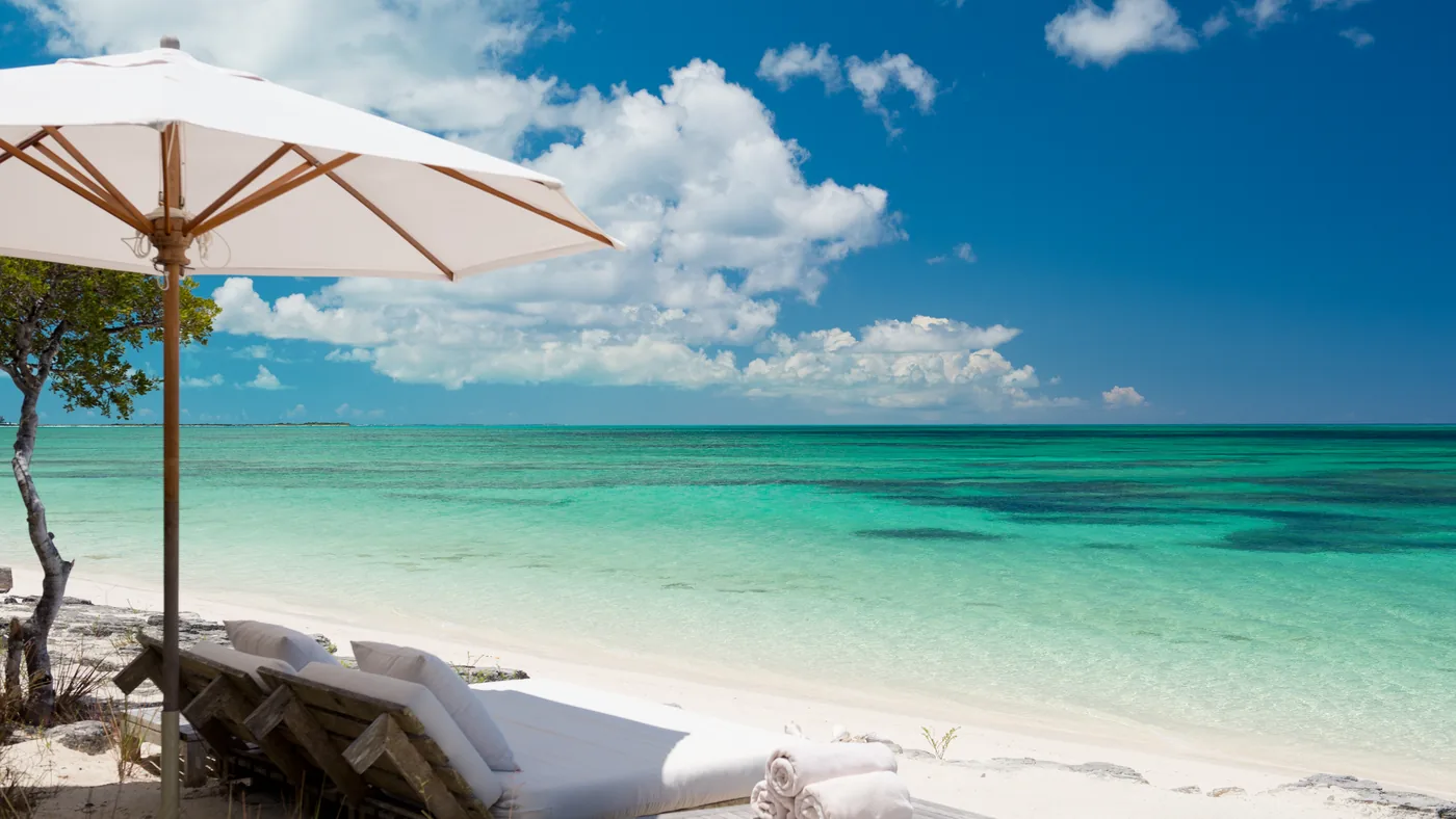 A cream umbrella and daybed loungers at the edge of clear turquoise water