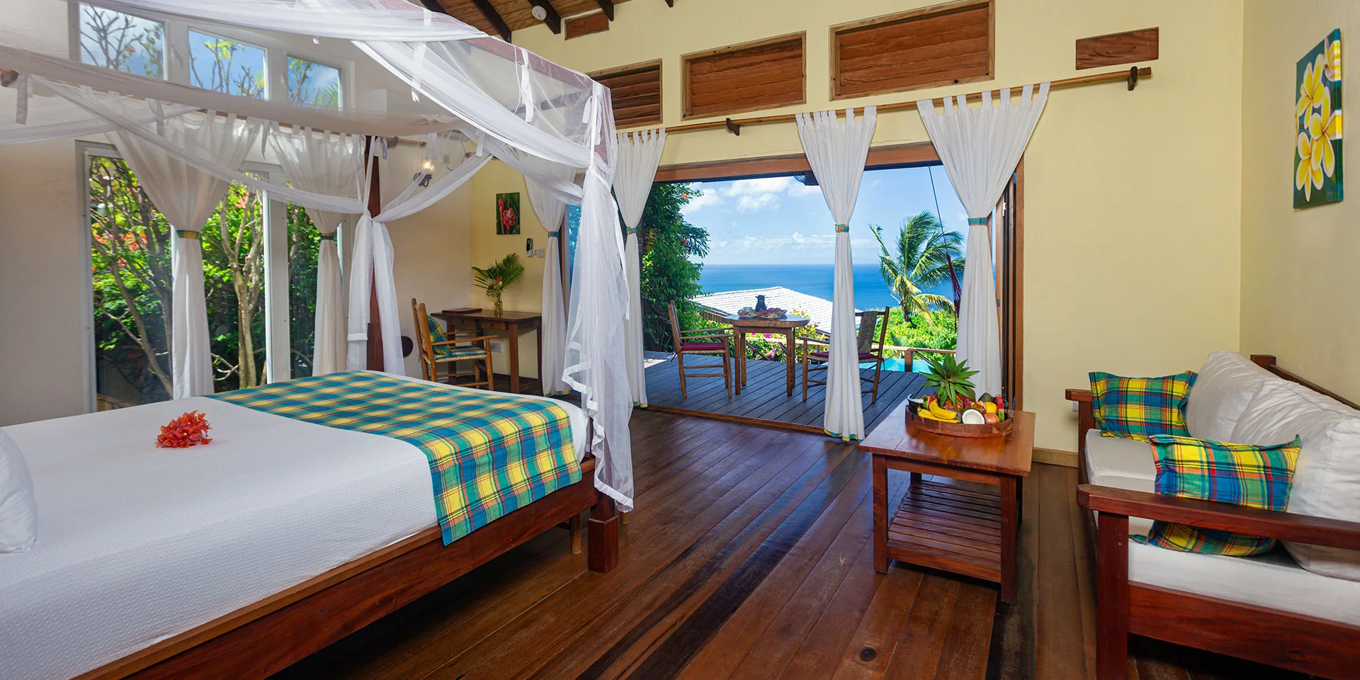 A canopied four-poster suite in blue plaid, doors open to a terrace over the Caribbean Sea